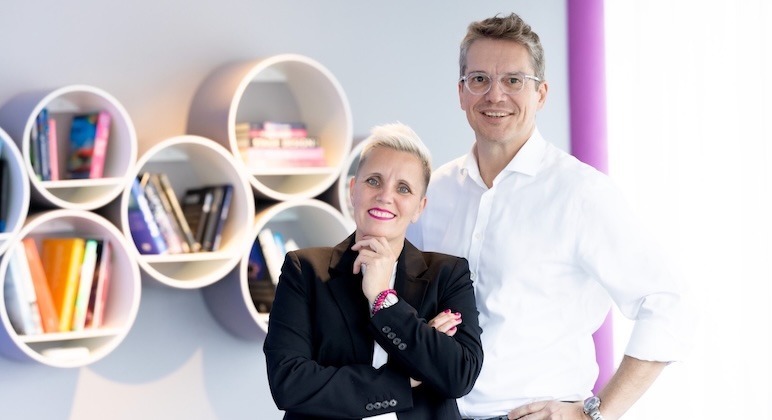 Rechtsanwältin Christiane Henneken, freundlich lächelnd, Arme verschränkt, linke Hand unter dem Kinn. Kopf leicht nach links geneigt. Schwarzen Blazer und weißes T-Shirt sowie pinkfarbenes Perlenarmband tragend. Dahinter stehend Rechtsanwalt Martin W. Schrenk. Freundlich lächelnd. Rundliche Brille mit durchsichtigem Rahmen und weißes Oberhemd ohne Krawatte tragend.