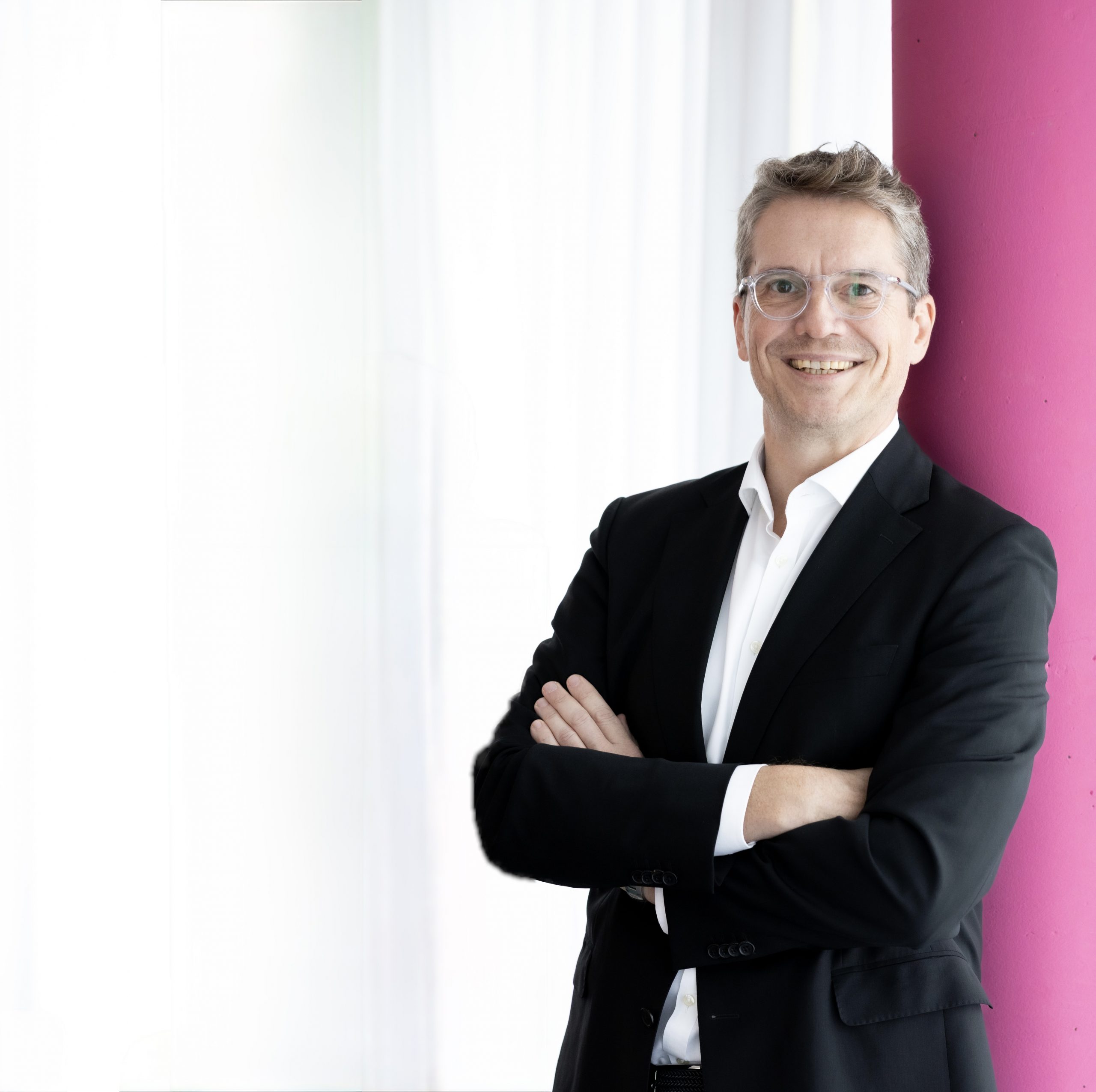 Rechtsanwalt Martin W. Schrenk an eine pinkfarbene Säule gelehnt. Freundlicher Blick. In schwarzem Sacko und weißem Oberhemd gekleidet. Er trägt Brille mit durchsichtigem Rahmen.