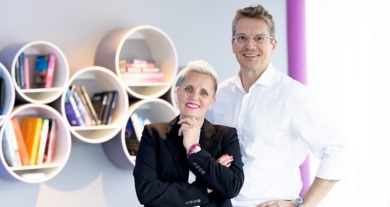 Rechtsanwältin Christiane Henneken, freundlich lächelnd, Arme verschränkt, linke Hand unter dem Kinn. Kopf leicht nach links geneigt. Schwarzen Blazer und weißes T-Shirt sowie pinkfarbenes Perlenarmband tragend. Dahinter stehend Rechtsanwalt Martin W. Schrenk. Freundlich lächelnd. Rundliche Brille mit durchsichtigem Rahmen und weißes Oberhemd ohne Krawatte tragend.